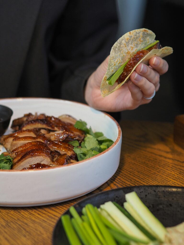 Close-up of a hand holding a duck wrap above a bowl of sliced duck with fresh greens.